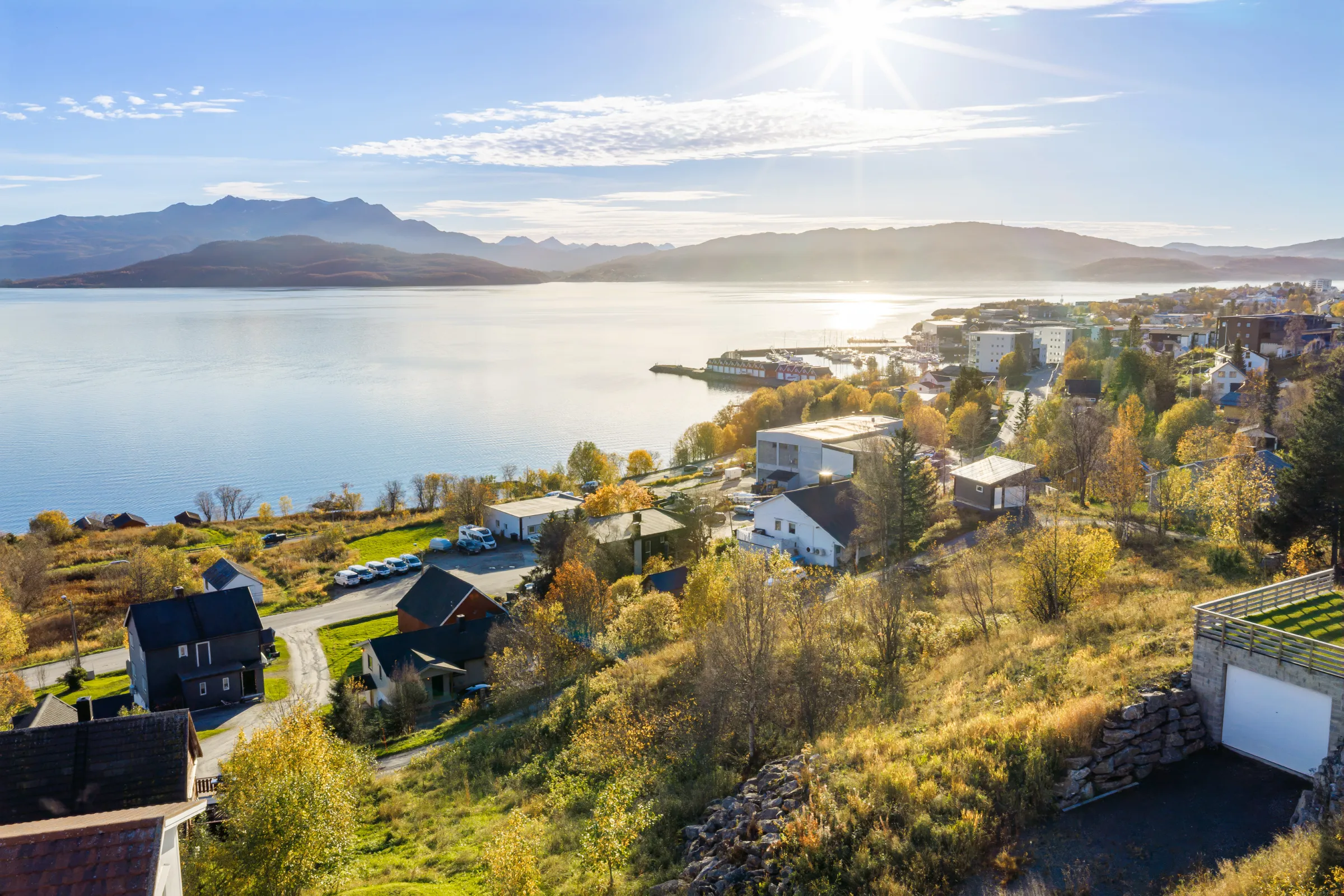 Dronebilde fra tomta mot sørvest: sjøen, småbåthavna og fjellene i ettermiddagssol.